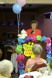 A photographic image of Mary Hunt Webb speaking in Cortez, Colorado, while a stuffed lamb rests on the podium).